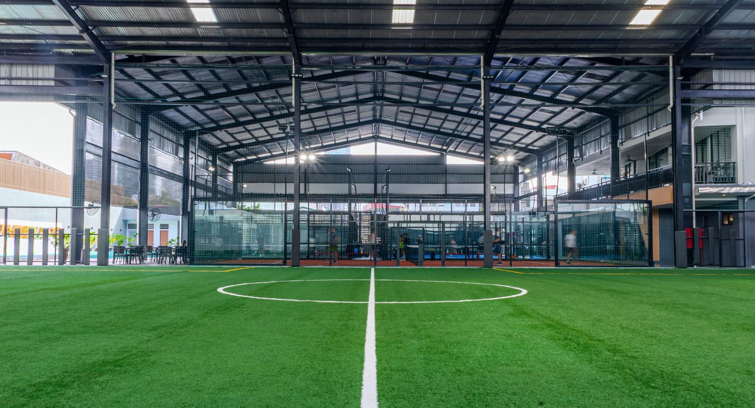 Indoor football pitch under a steel roof, with glass padel courts behind